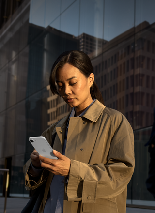 A person using a white iPhone 17e to continue work outside of an office building