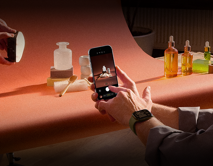 A photoshoot in a workplace setting, one person is using an iPhone to photograph products against a backdrop, one person is holding a light